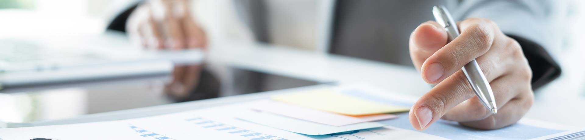 Close-up of a hand holding a silver pen over documents with colorful sticky notes on a desk.
