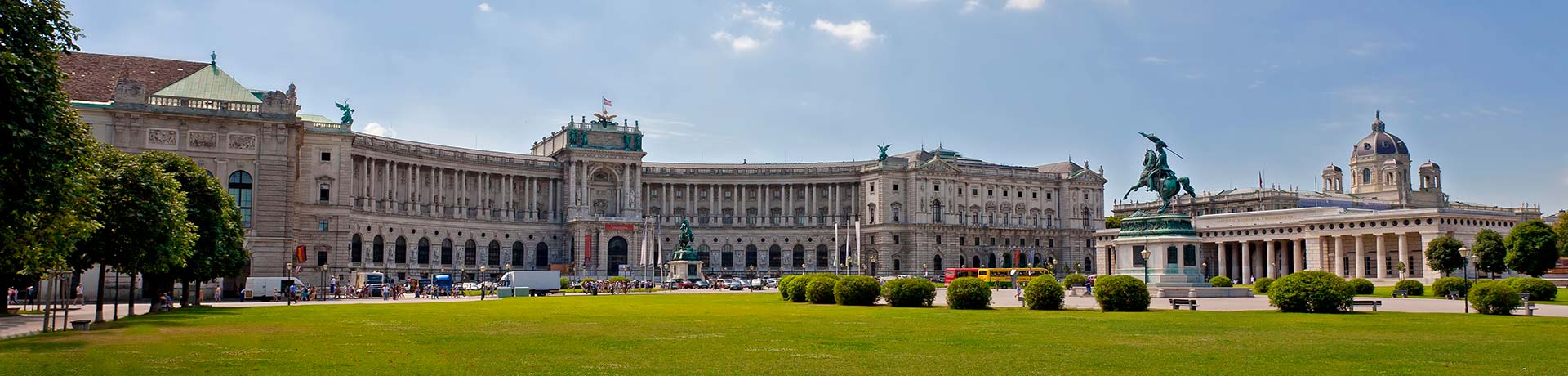 Wide view of a grand neoclassical palace with a large lawn and an equestrian statue in front, under a blue sky.
