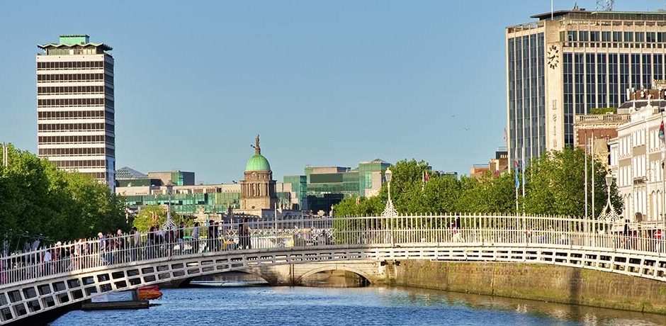 Wide view of a river with a white arched pedestrian bridge, green trees on the left, and red brick buildings lining the waterfront; people stroll along the promenade.