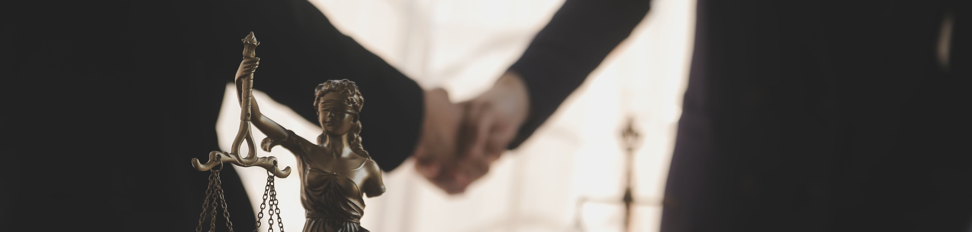 Bronze statue of Lady Justice holding scales in the foreground, with a blurred handshake in the background representing a legal agreement.